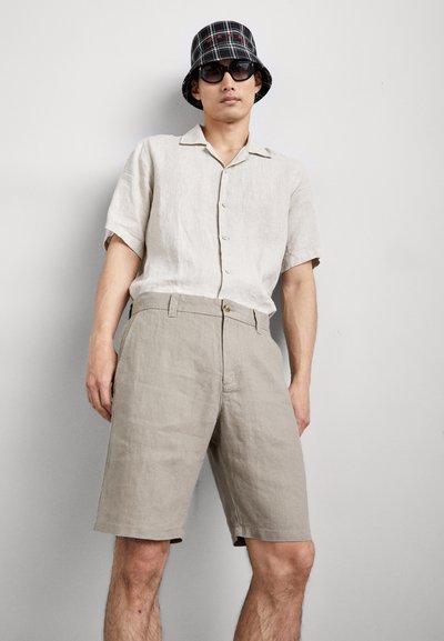 Une chemise en lin gris clair à manches courtes avec des boutons, associée à un short en lin beige et un chapeau seau à carreaux noirs. Des lunettes de soleil noires sont portées.