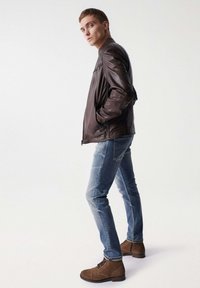 Braune Lederjacke mit Stehkragen, kombiniert mit hellblauen, verwaschenen Jeans und braunen Wildlederstiefeln. Das Model steht mit Händen in den Taschen.