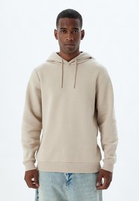 Sudadera beige con cordón y bolsillo tipo canguro. Presenta mangas largas, puños y cintura acanalados. Textura de tela suave.