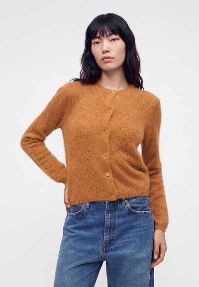 Femme aux longs cheveux foncés portant un cardigan marron boutonné et un jean bleu, debout une main sur la hanche devant un fond uni.