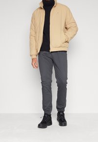 Hnědozlatá péřová bunda s elastickými manžetami, v kombinaci s tmavě šedými zúženými kalhotami a černými teniskami. Bunda m á zipový uzávěr a logo na detail. 