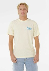 Blonde man met een lichtgele Rip Curl Board Bakery t-shirt en lichtblauwe jeans, staand tegen een witte achtergrond.