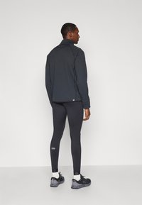 Jachetă athletică neagră cu guler înalt, colanți lungi negri, fitted, și adidași negri. Textură netedă, design minim, și detalii subtile ale logo-ului.