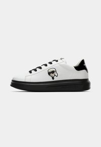 Białe skórzane sneakersy z czarnymi sznurowadłami i podeszwą, z logo z rysunkową twarzą po boku oraz czarnym akcentem na pięcie.