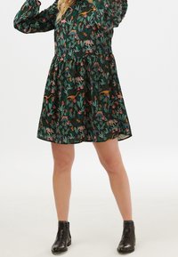 Vestido verde oscuro con estampado floral y de animales en rosa, naranja y azul. Mangas largas y falda acampanada. Combinado con botas negras hasta el tobillo.