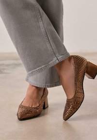 Chaussures à talons hauts tissées marron avec un bout carré et un talon bloc, complétées par un pantalon cropped gris clair dans un cadre neutre.