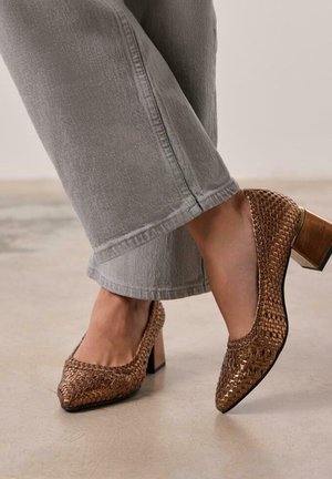 Chaussures à talons hauts tissées marron avec un bout carré et un talon bloc, complétées par un pantalon cropped gris clair dans un cadre neutre.