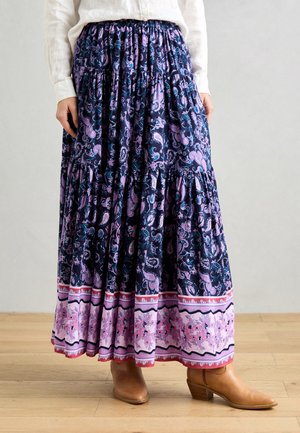 Jupe longue avec un motif paisley bleu foncé et violet, taille froncée, design à volants et un ourlet à motifs roses. Portée avec des bottes beiges.