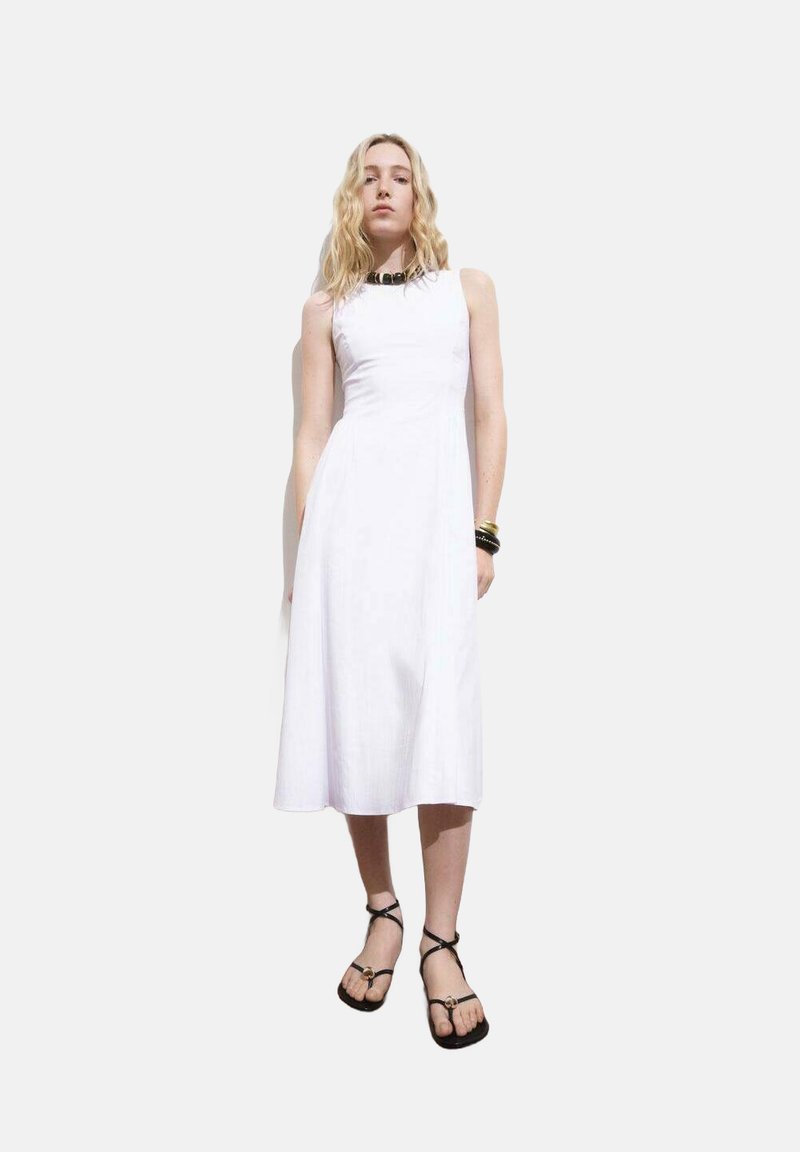 Robe midi blanche sans manches avec un corsage ajusté, une jupe fluide et une texture lisse. Associée à des sandales noires à lanières et des accessoires simples.