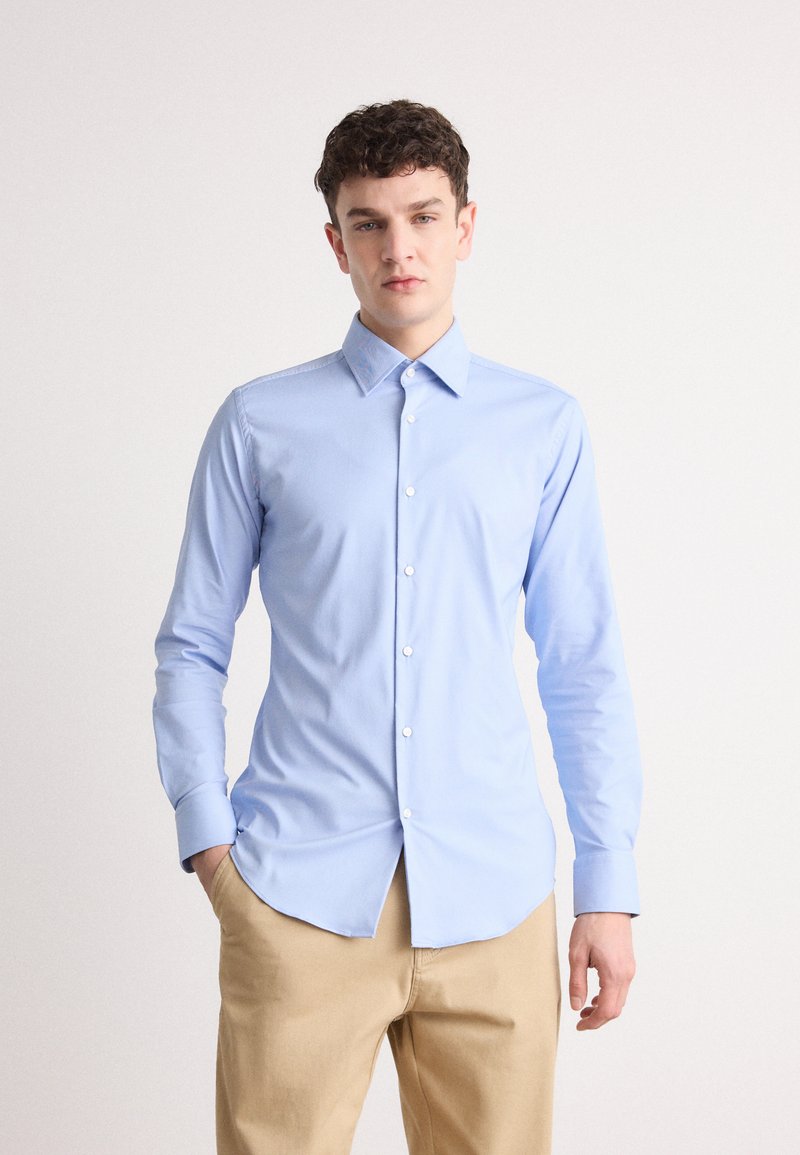 Camisa de manga longa azul clara com colarinho abotoado, design ajustado, textura de tecido suave e botões brancos. Usada com calças bege.