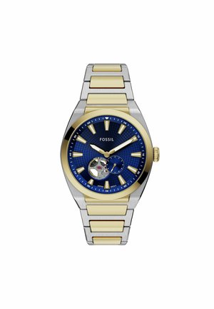 Reloj Fossil con un dial texturizado azul, acentos en tono dorado y una correa de acero inoxidable plateado. Presenta un movimiento esqueletado y marcadores de hora luminosos.