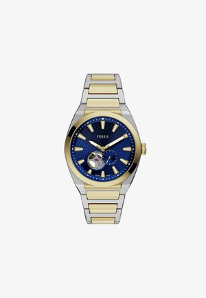 Montre Fossil avec un cadran texturé bleu, des accents dorés et un bracelet en acier inoxydable argenté. Présente un mouvement squelettique et des index d'heure lumineux.