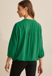 Blusa verde con una vestibilità morbida, maniche a 3/4 e dettagli arricciati sul retro. Tessuto morbido e leggero con una consistenza liscia.