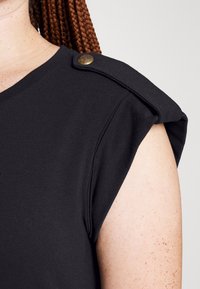 Černý textilní top má kulatý výstřih, strukturovaný design ramen a dekorativní mosazný knoflík na švu ramene.