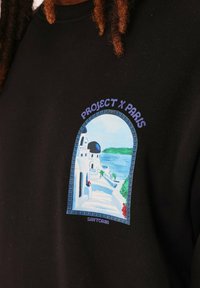 Sudadera negra con un diseño gráfico colorido de Santorini que presenta edificios azules y blancos, agua y plantas, etiquetada como "PROJECT X PARIS."