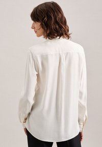 Weiße Bluse mit langen Ärmeln, glatter Textur, rückseitigem Faltendetail und abgerundetem Saum. Verfügt über geknöpfte Manschetten und einen klassischen Kragen.