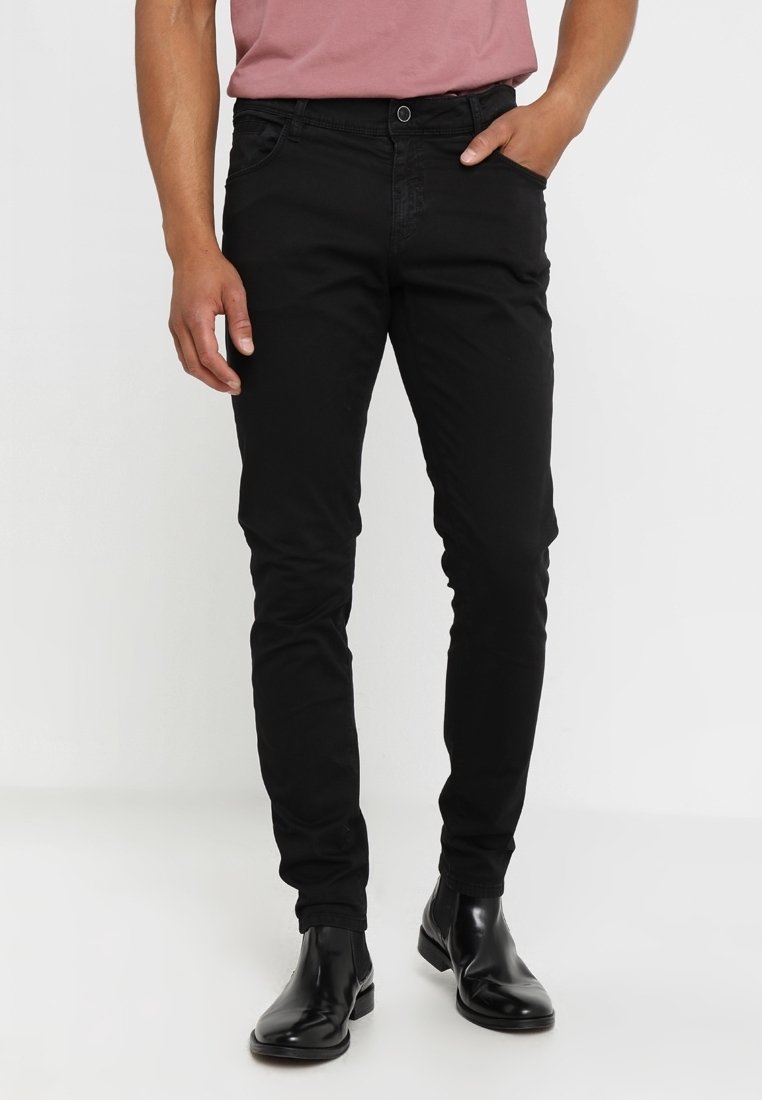Homme portant un pantalon noir slim et des bottines en cuir noir, debout avec une main dans la poche sur un fond uni.
