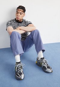 T-shirt rayé noir et blanc, pantalon violet clair, et chaussures de course grises avec des accents jaunes. Assis sur une surface moquettée bleue.