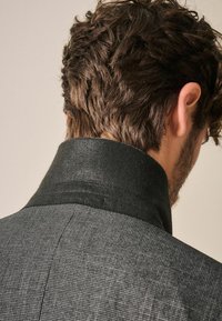 Blazer gris texturizado con un cuello de fieltro negro, que presenta un sutil patrón de cuadros y líneas limpias a lo largo de la espalda y los hombros.
