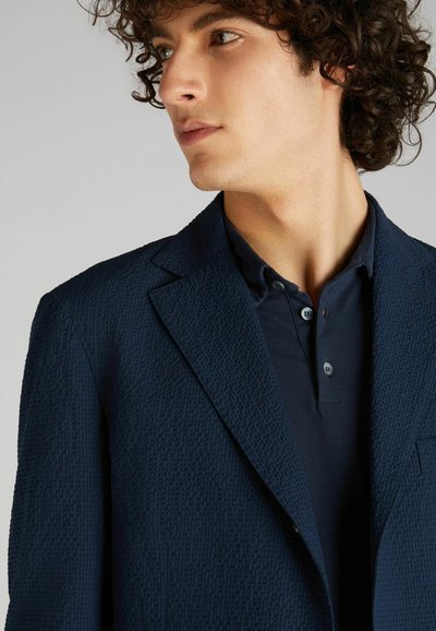 Granatowy marynarka o teksturowanej powierzchni, wykonana z węgińskiego materiału, z kołnierzem typu notch i zapięciem na dwa guziki. Noszona na dopasowanej, ciemnoniebieskiej koszulce polo.