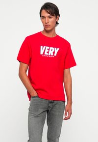 Vyras, vilkintis raudoną marškinėlį su užrašu "VERY CALVIN KLEIN", derinamas su pilkais džinsais, stovi prieš paprastą baltą foną.