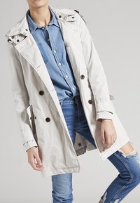 Trench coat gris claro con diseño de doble botonadura, gran solapa y bolsillos con botón, llevado sobre una camisa azul y unos jeans desgastados.