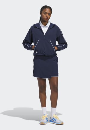 Joven de pie con las manos en los bolsillos, llevando una chaqueta con cremallera azul marino, falda a juego, polo blanco, calcetines blancos y zapatillas azules y amarillas.