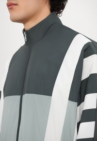 Grå og hvid zip-up jakke med høj krave, vandret farveblokering og et let, tekstureret stof.