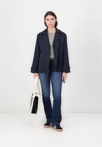 Marineblauwe, dubbelbreasted jas met grote knopen en schouderepauletten, gedragen over een lichtgrijze top en donkerblauwe jeans, terwijl ze een witte tas vasthoudt.