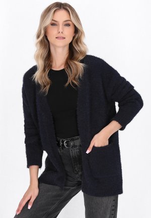 Navyblaue flauschige Strickjacke mit langen Ärmeln, offener Vorderseite und zwei vorderen Taschen, kombiniert mit einem schwarzen Oberteil und hochgeschlossenen grauen Jeans.