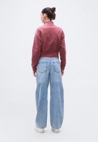 Μπορντό cropped φούτερ με ψηλό γιακά, συνδυασμένο με ανοιχτό μπλε τζιν παντελόνι με φαρδιά μπατζάκια. Λείο ύφασμα, άνετη εφαρμογή και ελάχιστες λεπτομέρειες ραφής.