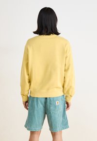 Sweatshirt jaune à manches longues avec des poignets et un ourlet côtelés, associé à des shorts teal. Tissu doux, design simple, sans motifs ni logos visibles.