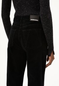 Zwarte corduroy broek met een getailleerd silhouet, voorzien van achterzakken en een merklabel op de tailleband. De textuur is ribbelig en zacht.