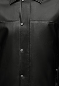 Μαύρο δερμάτινο μπουφάν με κουμπιά τύπου snap στο μπροστινό μέρος και ορατές ραφές στο στήθος και το γιακά.