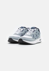 Sapatilhas atléticas cinzas com atacadores brancos e um upper texturizado. Apresenta um logótipo em azul e uma entressola branca em contraste.