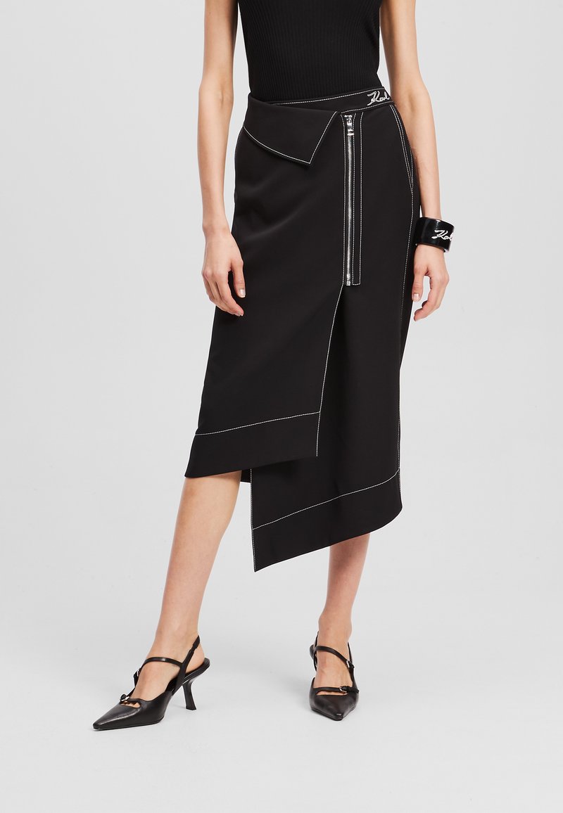 Zwarte asymmetrische rok met contrasterende witte stiksels, voorritsdetail en een diagonale zoom. Gecombineerd met zwarte slingback hakken.