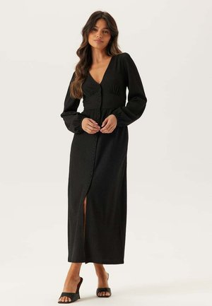 Kvinde iført en lang sort kjole med knapper, lange ærmer og en frontslids, stående mod en ensfarvet off-white baggrund.