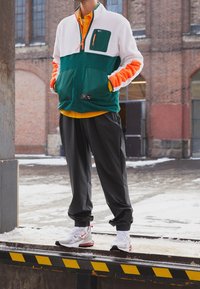 Grön och vit zip-up fleecejacka med orange ärmar och svarta byxor; har en bröstficka och logodetaljer. Bärs med vita och röda sneakers.