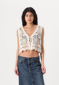 Biele háčkované crop top s kruhovými vzormi, V-krkom a detailom na zaväzovanie vpredu, skombinovaný s džínsovou sukňou s vysokým pásom.