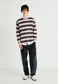 Jeune homme portant des lunettes de soleil orange, une chemise à manches longues rayée rose et noire, un t-shirt blanc en dessous, un pantalon noir et des baskets blanches, se tenant contre un fond blanc.