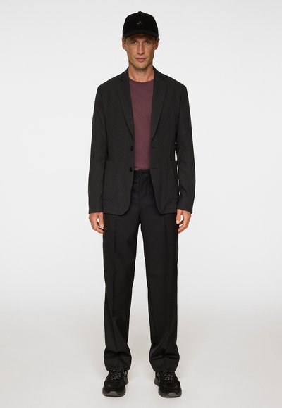 Blazer grigio scuro sopra una camicia bordeaux, abbinato a pantaloni neri e sneakers nere. Un cappellino nero completa il look generale.