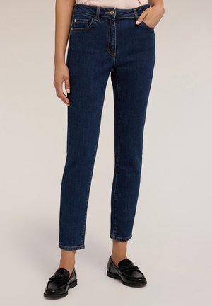 Vrouw in donkerblauwe slim-fit jeans en zwarte loafers, staande met één hand in de zak tegen een neutrale achtergrond.