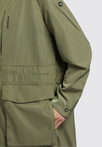 Olivgrön lätt jacka med huva, stora fickor och en jämn yta. Svart logotypdetalj på axeln.