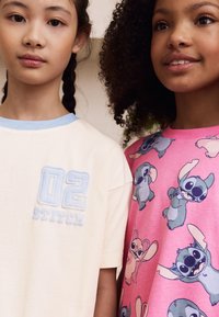 Zwei Mädchen tragen T-Shirts: eines in Creme mit blauer Einfassung und einem "02 STITCH"-Aufdruck, das andere in Pink mit einem Muster aus Stitch-Figuren.