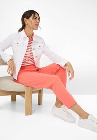 Witte spijkerjacket met gouden knopen, rood-wit gestreept poloshirt, koraalkleurige wijde broek en witte sneakers. Zittend op een lichthouten stoel.
