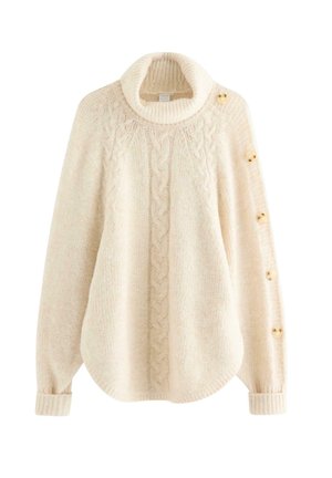 Junge Frau mit langen blonden Haaren, die einen übergroßen cremefarbenen Rollkragenpullover mit Knöpfen an den Ärmeln, graue Jeans und beige Sneakers trägt.
