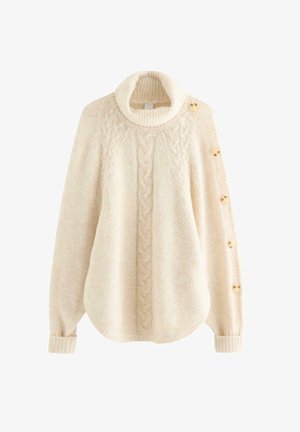 Junge Frau mit langen blonden Haaren, die einen übergroßen cremefarbenen Rollkragenpullover mit Knöpfen an den Ärmeln, graue Jeans und beige Sneakers trägt.