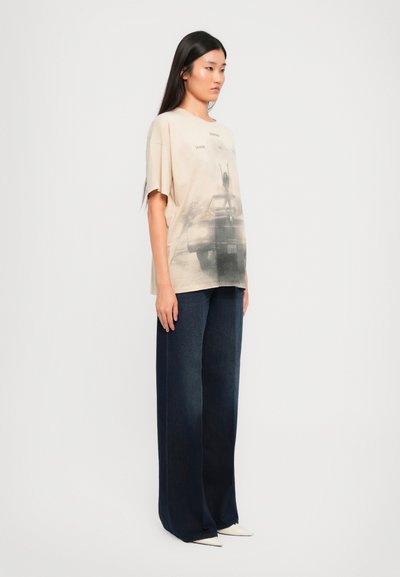 Camiseta gráfica beige con un estampado de coche desvaído, combinada con unos pantalones anchos oscuros y zapatos blancos de punta. Textura de algodón suave.