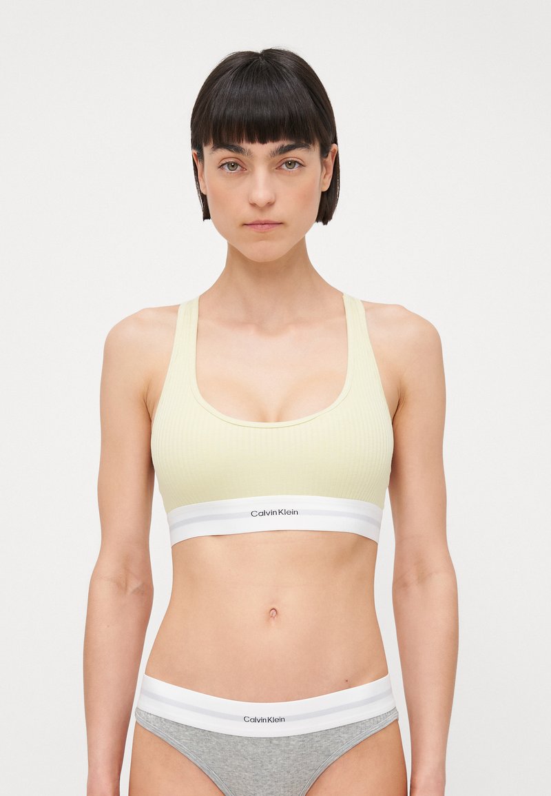 Lys gul, ribbet sports-BH med bred, hvit elastisk kant som har logo. Sammen med grå undertøy, også med hvit kant.