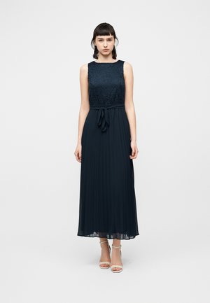 Kvinde iført en navyblå, ærmeløs kjole med blonder på overdelen, bundet talje, plisseret skørt og hvide sko med ankelrem, stående foran en ensfarvet baggrund.